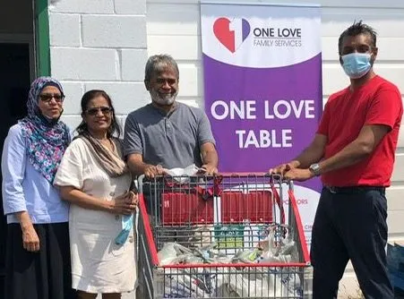 Volunteers with a shopping cart full of donated groceries beside the One Love Table banner